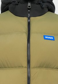 Puffer jacket in green and black with a zip front, featuring a blue logo patch and drawstring hood. Textured, insulated design.