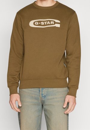Person trägt einen braunen Langarm-Sweatshirt mit dem "G-STAR"-Logo in Beige auf der Brust und hellblaue Jeans.