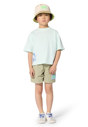 Light green cropped t-shirt with blue graphic, beige bucket hat with green accent, khaki shorts with patch, and white shoes with blue laces.