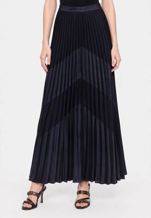 Vrouw draagt een lange zwarte geplooide rok met chevronpaneelpatronen, gecombineerd met zwarte sandalen met hoge hakken en studdetails.