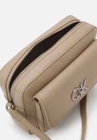 Beżowa skórzana torba na ramię z srebrnym zamkiem, regulowanym paskiem, przednią kieszenią z klapką oraz srebrnym logo CK na białym tle.