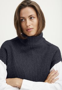 Dicker Strick-Turtleneck-Pullover in dunklem Marineblau, mit einem strukturierten Muster und kombiniert mit einem weißen Hemd darunter.