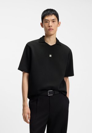 Junger Mann trägt ein schwarzes kurzärmeliges Poloshirt mit kleinem Logo auf der Brust und schwarze Hose mit Gürtel vor weißem Hintergrund.