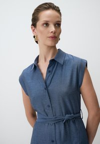 Ein blaues kurzärmeliges Hemdkleid aus weichem Stoff mit einer durchgehenden Knopfleiste und einem gebundenen Taillengürtel. Es hat einen klassischen Kragen und eine glatte Textur.