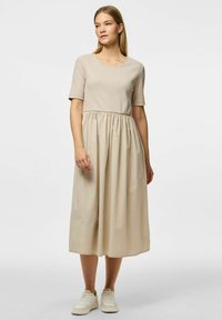 Bež haljina od mješavine pamuka s kratkim rukavima, skupljenim po struku i suknjom koja se širi do koljena. Uparena s bijelim tenisicama.