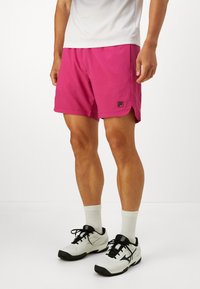 Man wearing bright pink athletic shorts, white crew socks, and white and black sports shoes standing against a plain white background.