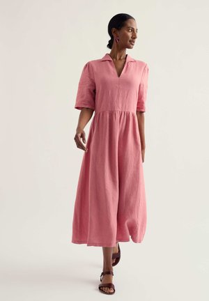 Žena kráča v ružových šatách strednej dĺžky, voľného strihu s krátkymi rukávmi, ktoré kombinuje s tmavými sandálmi a purpurovými visiacími náušnicami.