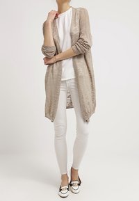 Long, beige-knit cardigan over a white textured top, paired with white leggings and black-and-white loafers featuring gold accents.