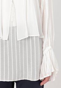Blusa bianca con un design fluido, caratterizzata da maniche a campana e un motivo a righe testurizzato sotto. Tessuto morbido con un aspetto leggero.