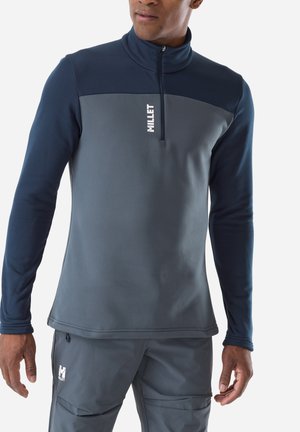 Mann trägt ein zweifarbiges blau-graues Millet Langarm-Quarter-Zip-Oberteil und passende Hose, steht mit locker an den Seiten hängenden Armen.