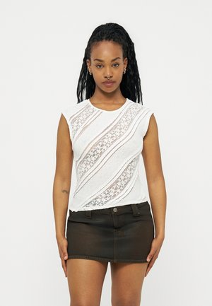 Νέα γυναίκα με μακριά πλεξούδα φοράει λευκό αμάνικο δαντελένιο τοπ και μαύρη μίνι φούστα από τζιν, στέκεται μπροστά σε απλό φόντο.