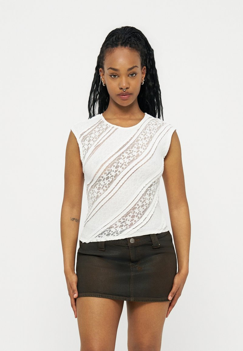 Νέα γυναίκα με μακριά πλεξούδα φοράει λευκό αμάνικο δαντελένιο τοπ και μαύρη μίνι φούστα από τζιν, στέκεται μπροστά σε απλό φόντο.