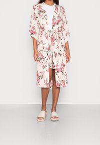 Długi, kwiatowy kimono w kremowym kolorze z różowymi kwiatami i zielonymi liśćmi. Lekki, wiązany w talii, z szerokimi rękawami, zestawiony z beżowymi klapkami.