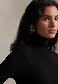 Pulover negru cu guler înalt și model cu dungi, croială strânsă. Dispune de mâneci lungi și un guler înalt. Modelul are păr ondulat și fundal neutru.