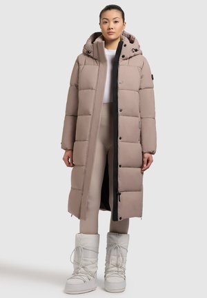 Lange, gewatteerde jas in lichtbeige met capuchon, zwarte rits en knopen. Heeft een geborduurd ontwerp en zijzakken. Gecombineerd met witte laarzen.
