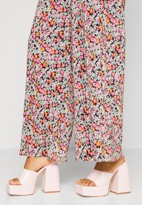 Sandales à plateforme roses avec une finition brillante, large bride et talon en bloc géométrique, associées à un pantalon large à motifs floraux.