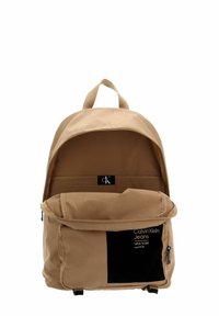 Sac à dos beige en tissu avec fermeture éclair, doté d'une poche avant, d'un logo marqué en évidence et d'une poignée supérieure pour le transport.