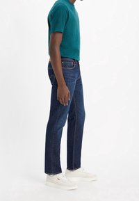 Teal crew-neck t-shirt åker ihop med mörkblå denimjeans. Jeansen har orangea sömmar och rak ben-design. Vita sneakers fulländar looken.