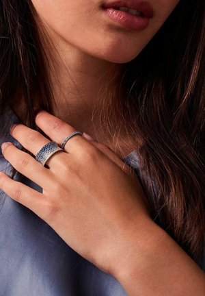 Silverringar med blå detaljer, bestående av en bred ring med strukturerad yta och en smalare ring, som bärs på en avslappnad hand mot huden.