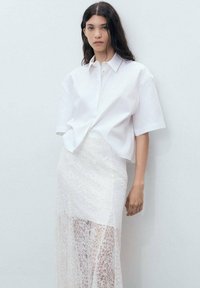 Cămașă albă cu mâneci scurte și guler, asociată cu o fustă din dantelă transparentă, având modele florale intricate și un design stratificat.
