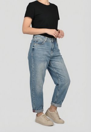 Personne portant un jean bleu clair retroussé, un T-shirt noir à manches courtes et des baskets beiges, debout devant un fond clair uni.