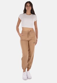 Pantaloni jogger beige con polsini elasticizzati, vita regolabile con cordino e tasche laterali; abbinati a un top corto bianco a maniche corte.