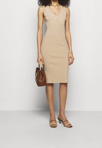 Femme portant une robe en maille beige sans manches, tenant un sac à main marron avec une poignée tissée, portant des talons à lanières bicolores et des bijoux en or.