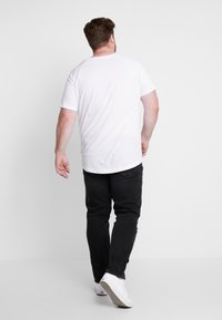 Vit kortärmad t-shirt med rund halsringning, i kombination med svarta jeans och vita sneakers, sedd bakifrån mot en enkel bakgrund.