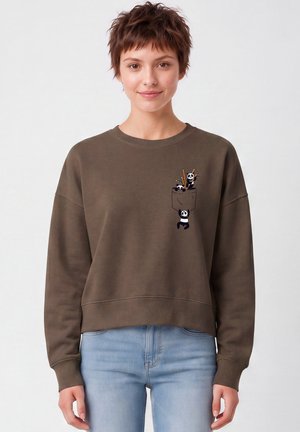 Junge Frau trägt braunen Sweatshirt mit Tasche, auf der drei verspielte Pandas mit Bambusstangen dargestellt sind, kombiniert mit hellblauen Jeans.
