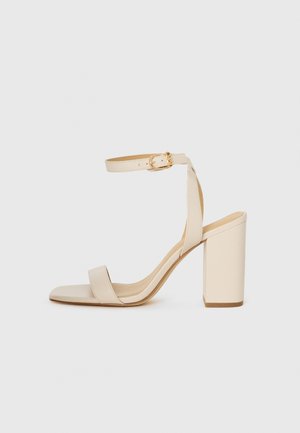 Sandal con tacco alto beige con punta quadrata, sottile cinturino alla caviglia e tacco a blocco. Texture di pelle liscia con dettaglio della fibbia dorata.
