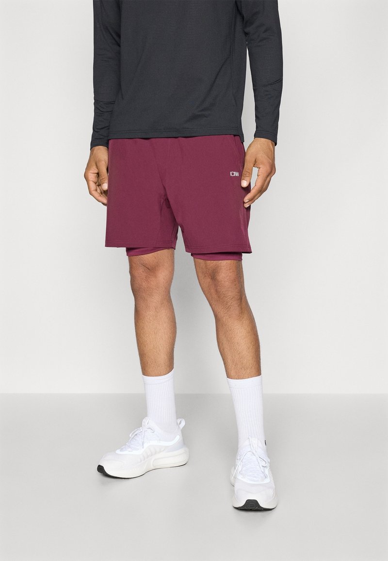 Homme portant un short de sport marron, un t-shirt noir à manches longues, des chaussettes hautes blanches et des chaussures de course blanches, debout devant un fond uni.