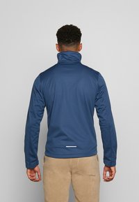 Veste bleu marine en tissu lisse et extensible ; elle présente un col montant et un accent réfléchissant à l'arrière près de l'ourlet.