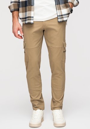 Mężczyzna w beżowych spodniach typu cargo, białych trampkach, białej koszuli oraz koszuli w kratę, z rękami w kieszeniach, stoi na neutralnym tle.