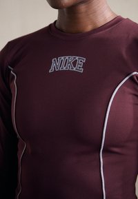 Viininpunainen urheilupaita pitkillä hihoilla, jossa on valkoinen Nike-logo ja erottuva valkoinen tikkaus saumoissa. Sileä pinta.
