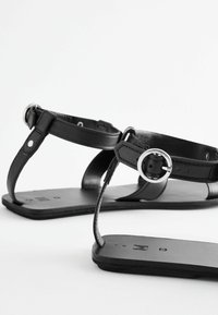 Černé kožené sandály s plochou podrážkou, s nastavitelnými popruhy a kruhovými kovovými akcenty. Minimalistický design a hladký povrch.