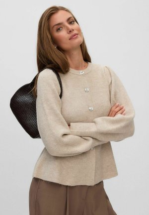 Femme aux longs cheveux châtain clair portant un pull beige boutonné et un pantalon marron, portant un sac à épaule noir tissé, bras croisés.
