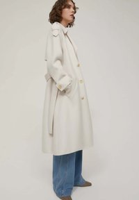 Lungo cappotto crema con grandi bottoni, tasche laterali e spalline. Abbinato a jeans a gamba larga blu chiaro e calzature beige.