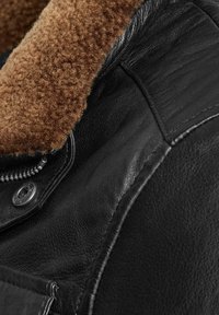 Zwarte leren jacket met een bruin fluffy kraag. Details omvatten een metalen rits en knopen, waarmee de textuur en het materiaalkontrast worden benadrukt.