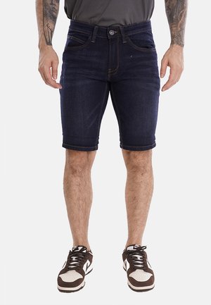 Shorts di jeans scuri con una vestibilità slim, dotati di chiusura con bottone e dettagli cuciti. Abbinati a scarpe da ginnastica marroni e bianche.