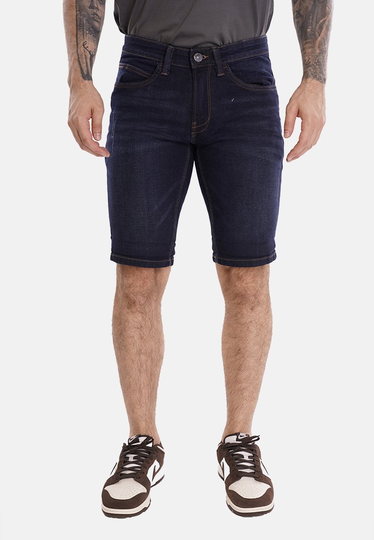 Shorts di jeans scuri con una vestibilità slim, dotati di chiusura con bottone e dettagli cuciti. Abbinati a scarpe da ginnastica marroni e bianche.