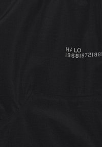 Tessuto nero con cucitura ricamata e testo stampato bianco "HALO 19681972198" parzialmente visibile nell'angolo in alto a destra.