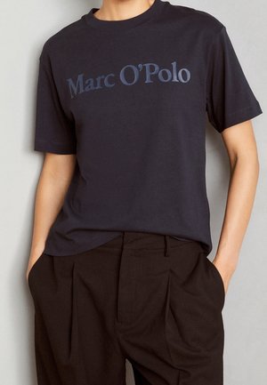 Námořnická modrá bavlněná trička s krátkým rukávem, s logem "Marc O'Polo" v kontrastní modré barvě. Nošeno s černými sklady kalhotami.