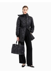 Giacca puffer nera trapuntata con cappuccio e vita regolabile, abbinata a una borsa a spalla nera trapuntata e pantaloni neri a gamba larga.