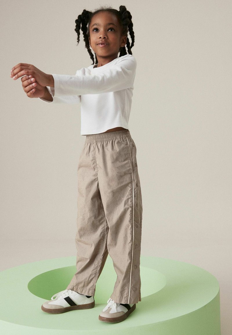 Haut crop en blanc à manches longues, pantalon beige à taille élastique avec des accents de boutons sur les côtés, et baskets blanches avec un détail de rayures noires.