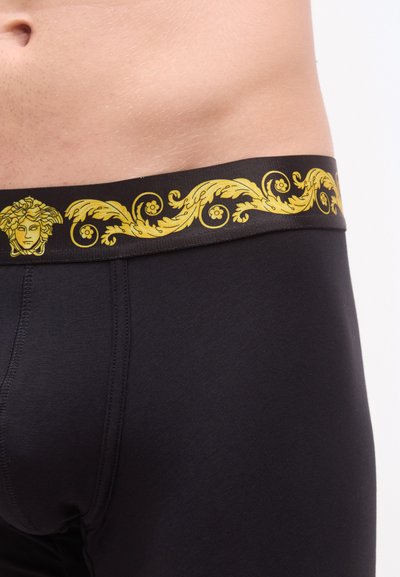 Μαύρα ανδρικά εσώρουχα boxer με χρυσή ελαστική ζώνη μέσης, διακοσμημένη με κλασικό σχέδιο που περιλαμβάνει λουλουδένια μοτίβα και μια λεπτομέρεια κεφαλής.