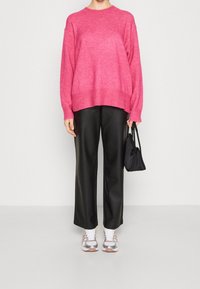 Osoba w jasnoróżowym swetrze z dzianiny, czarnych szerokich spodniach, białych i różowych trampkach, trzymająca małą czarną torebkę.