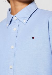 Person, die ein hellblaues Hemd mit Knopfleiste und einem kleinen rot-weiß-schwarzen Flaggenlogo auf der Brust trägt.