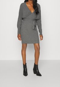 Robe portefeuille gris en tricot avec des manches longues et un lien à la taille, associée à des bottines noires à talon carré.