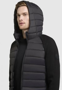 Piumino nero con cappuccio alto trapuntato e chiusura con zip. Presenta maniche in maglia nera a contrasto e un logo circolare sulla spalla.