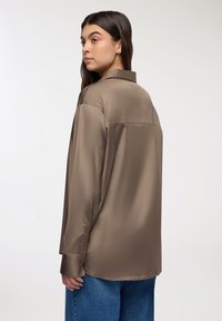 Camicia di satin marrone con colletto e maniche lunghe, caratterizzata da una vestibilità rilassata e un leggero lucido. Indossata con pantaloni in denim blu.